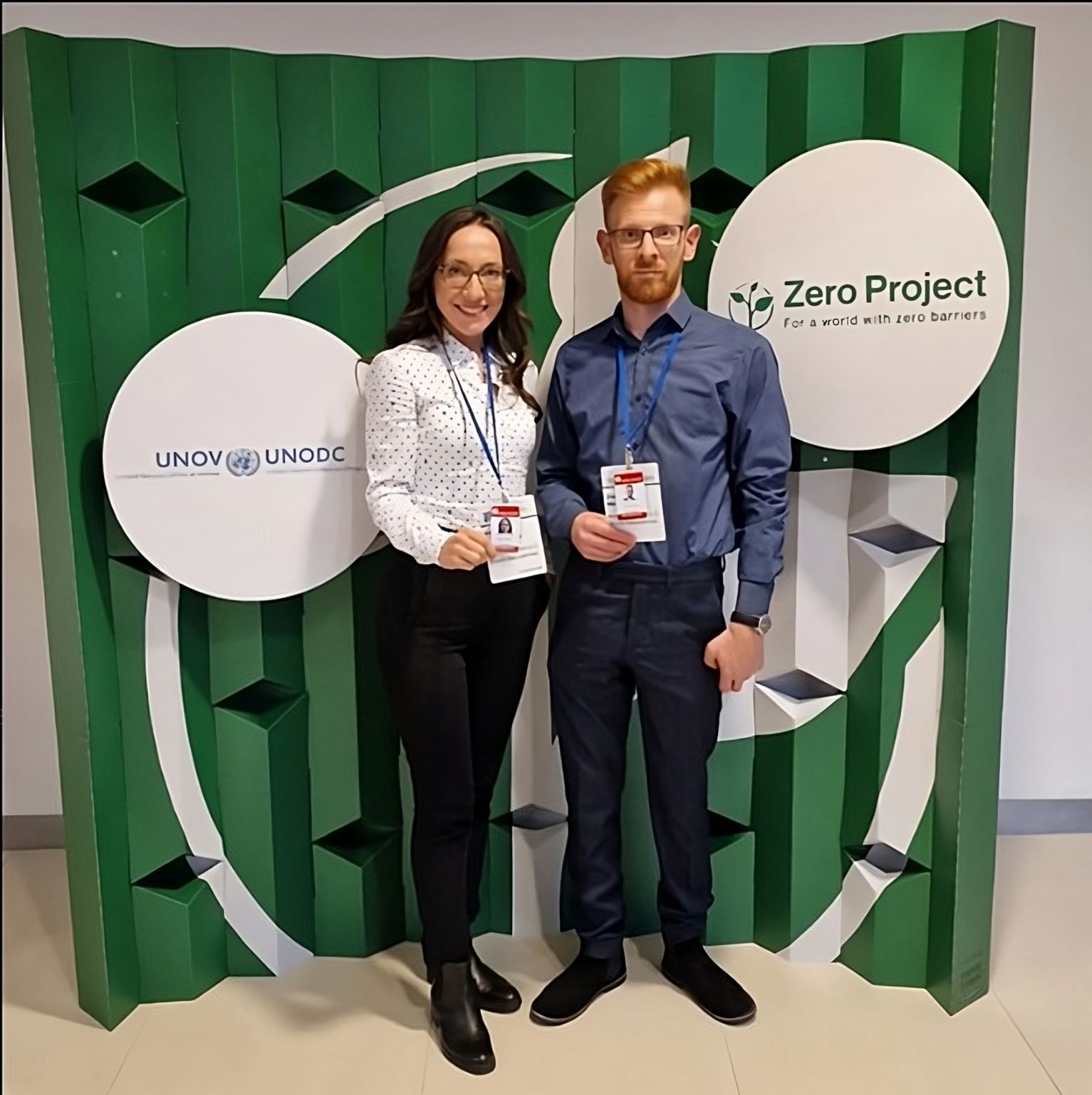 The CEO and founder Elizabeta with the digital marketer Dejan. 
The CEO has long wavy black hair. She is wearing glasses, white shirt with dots and black pants.
The digital marketer is ginger. Has a short hair and beard. He wears glasses, blue shirt and navy blue pants.

The background is a green screen with the logos of UNOV - UNODC and Zero Project.  
