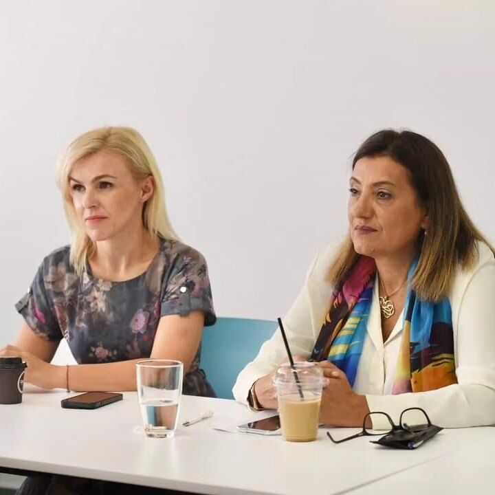 In this picture are Jovanka Trencevska, а Minister of Labor and Social Affairs and her assistant on the right. The minister wears a white shirt with a colourful scarf. She has brown long hair. 
The assistant wears a black T-shirt with flowers.